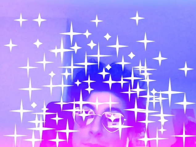 A white person with short dark curly hair and pink heart shaped glasses tilts his head. We have added 4-pointed angular white sparkles of varying sizes all over the photo. We also see a sparkle placed over each lens of the person's heart shaped glasses